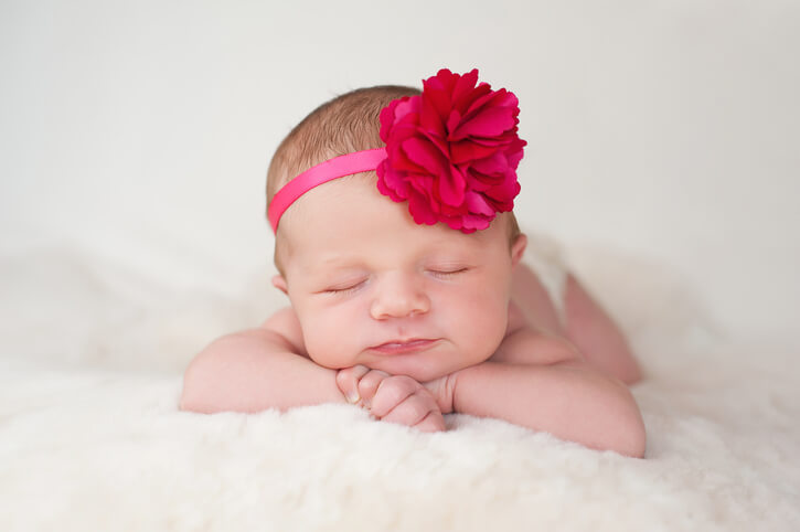 A portrait of a beautiful newborn baby girl wearing a hot pink flower headband. She is sleeping on a cream colored sheepskin rug.