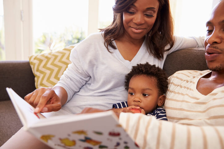 Parents And Son Reading Book On Sofa At Home Together