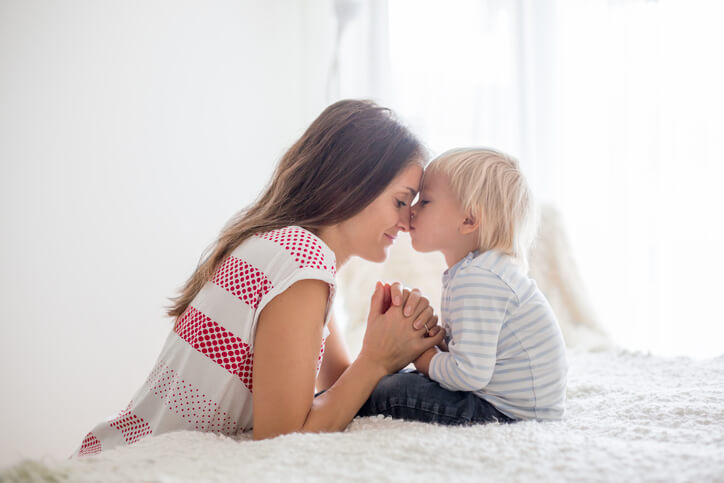 Mother and toddler son, praying at home, sitting on bed in bedroom